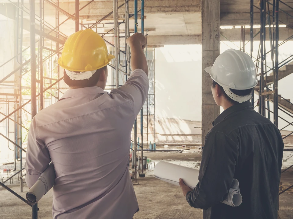 Chantier de construction intérieur avec deux professionnels du bâtiment portant casques de sécurité jaune et blanc, consultant des plans architecturaux